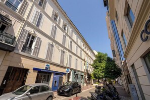 A street view with a building featuring a blue sign for a bank, parked cars, and a tree. - La Mansarde du Bivouac, cocon sous les toits (Cannes)