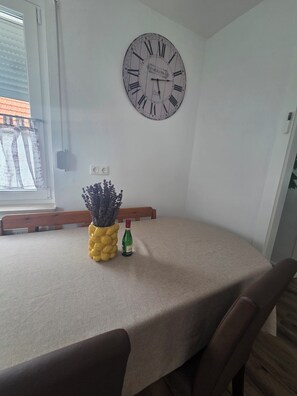 Zona pranzo con orologio rotondo a muro, tavolo con tovaglia beige, una bottiglia di vino e una pianta decorativa.