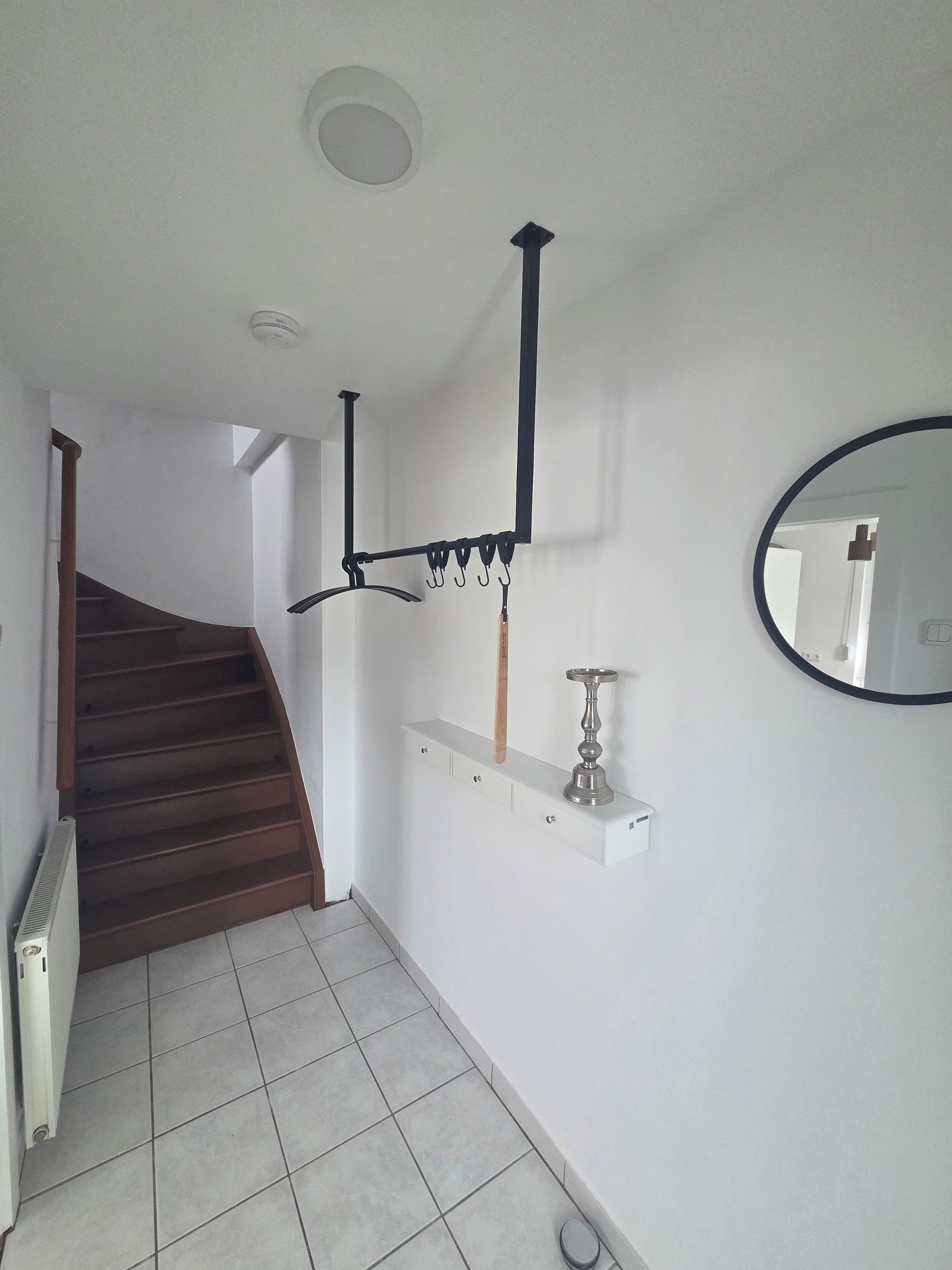 A staircase leading to a landing with a white wall, a black metal rack with hooks, a small shelf with a candle, and a round mirror.