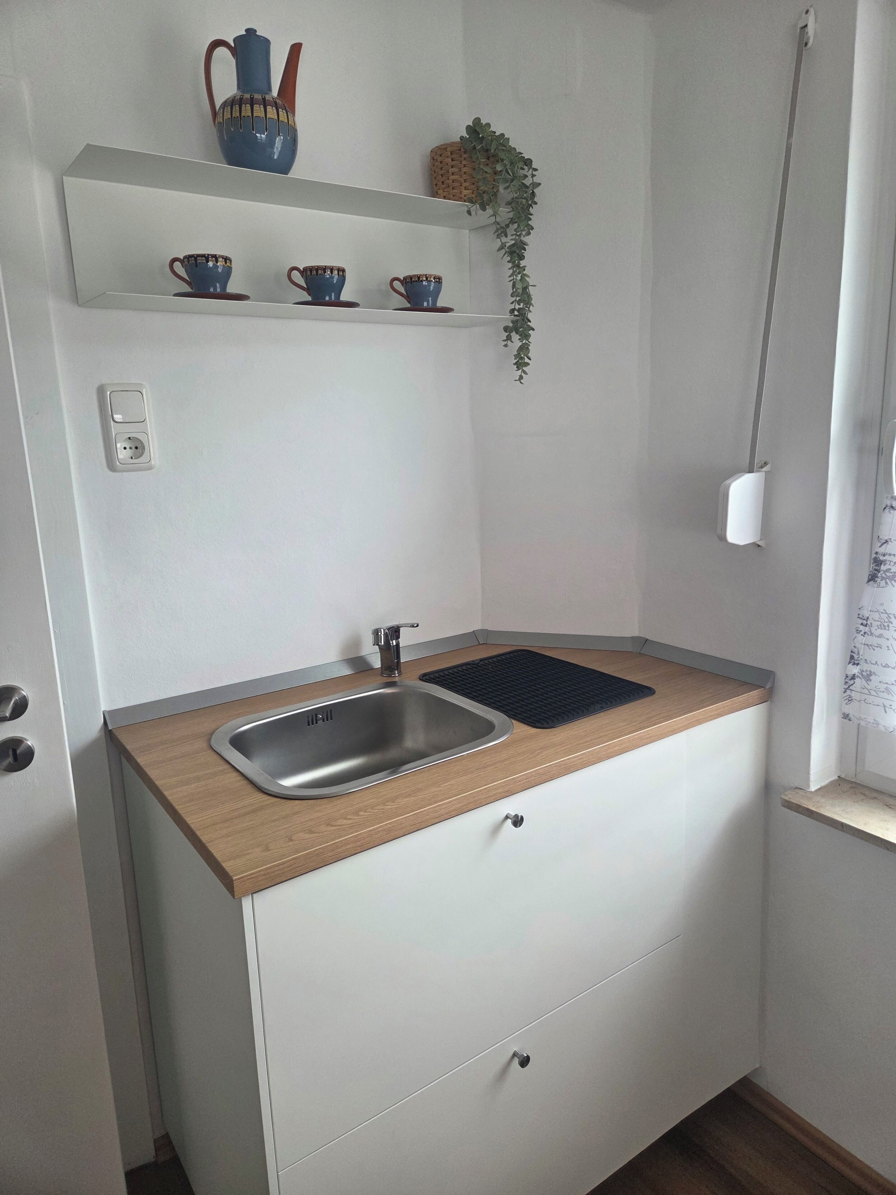A compact kitchen with a sink, wooden countertop, and a shelf above holding cups and a plant.