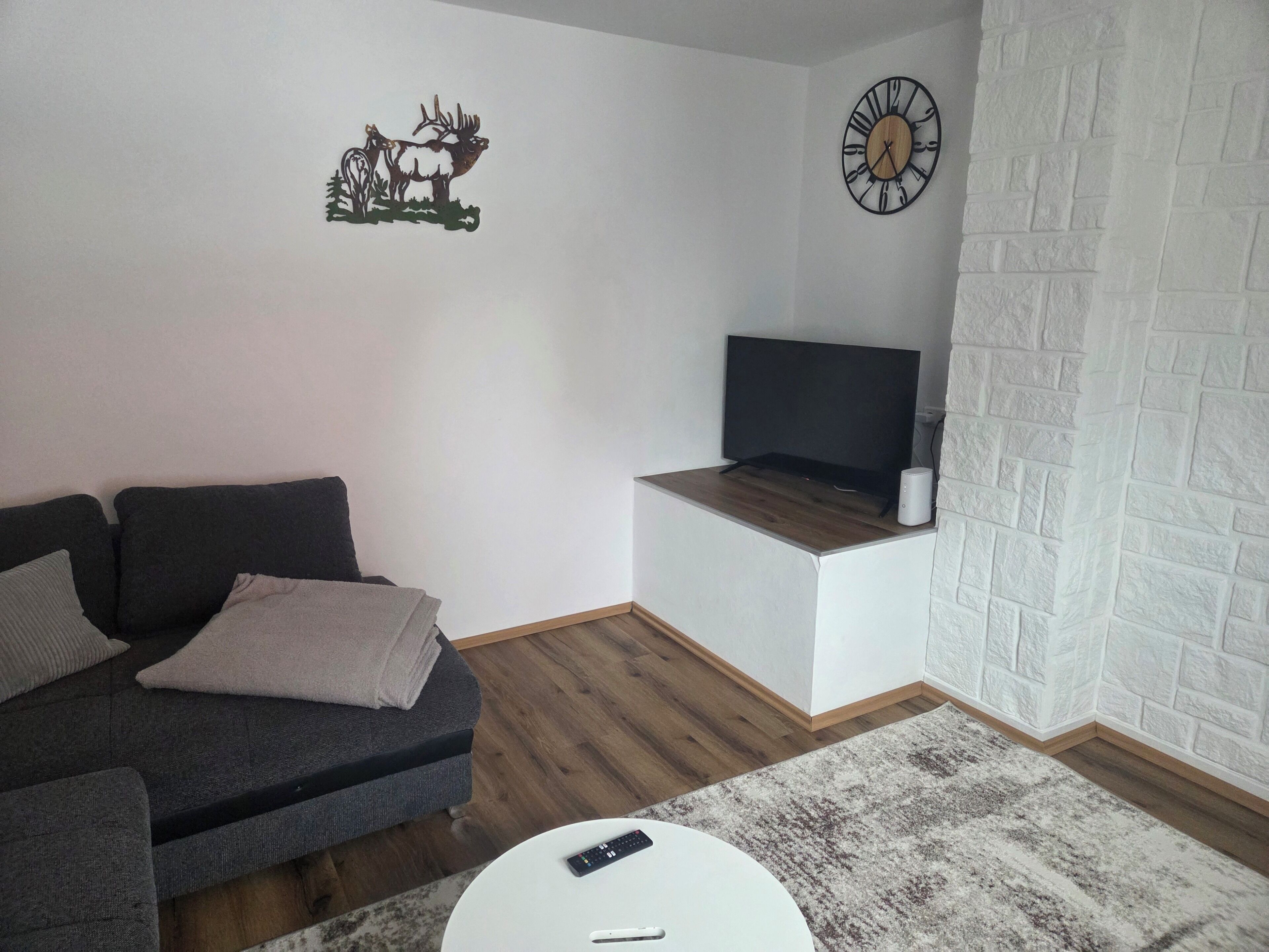 A living room with a sofa, a coffee table, a flat-screen TV, a clock, and a decorative wall piece.