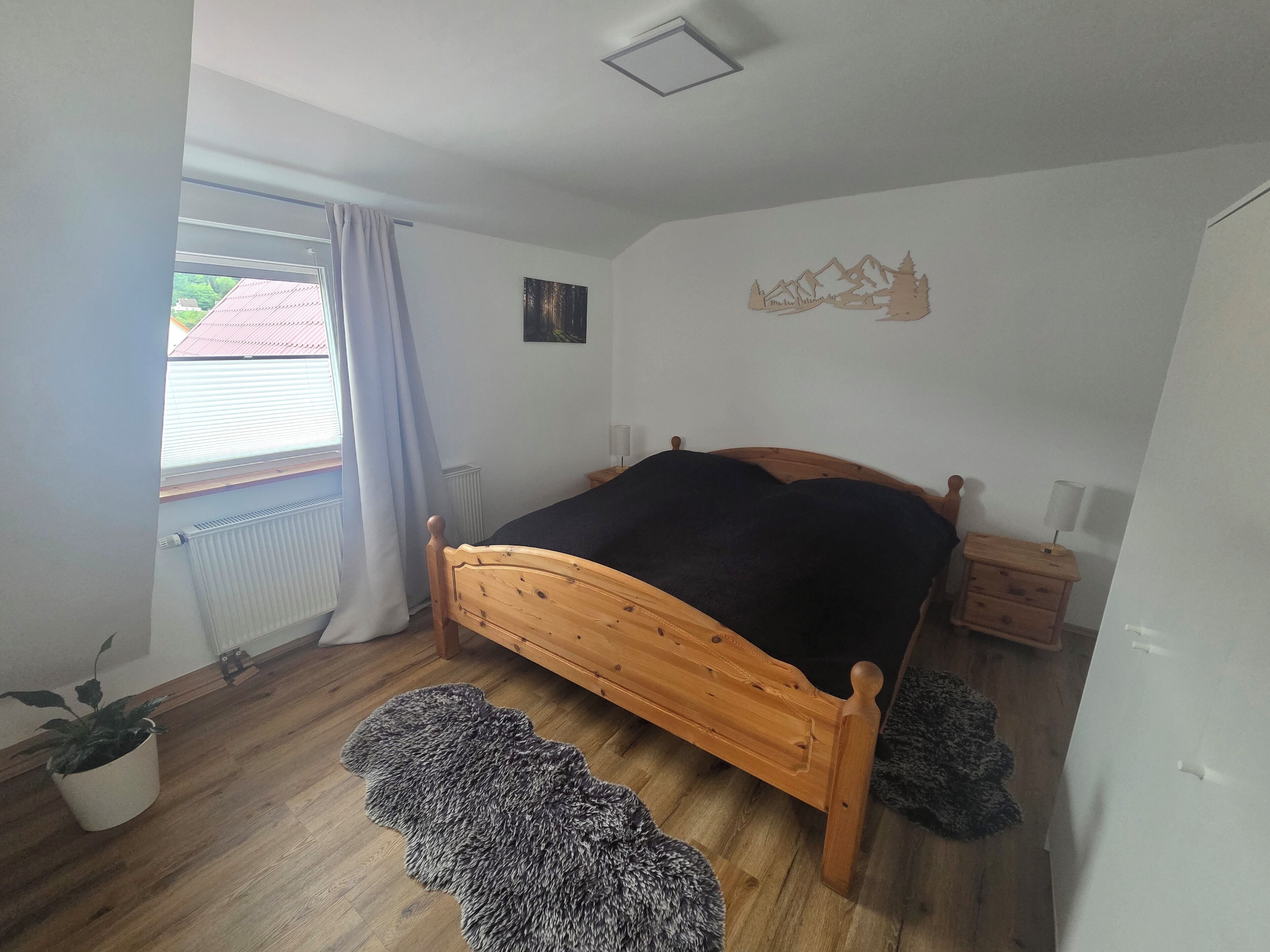 A bedroom with a wooden bed, a radiator, a window with curtains, a wall-mounted picture, and a small bedside table with a lamp.