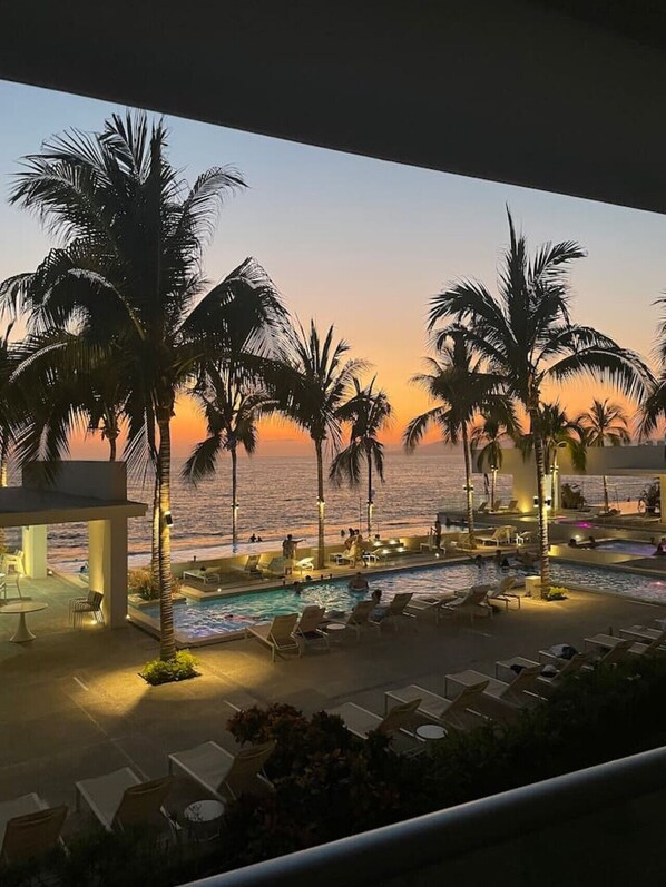 Poolbereich eines Resorts mit Palmen und Blick auf das Meer bei Sonnenuntergang.