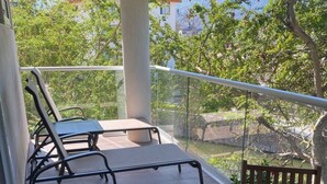 A wooden table and chairs on a balcony with a glass railing, overlooking a natural landscape with trees and a mountain in the background. - Luxury Beachfront | Corner Condo | Amazing Pools (Puerto Vallarta)