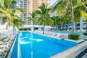 Ein Swimmingpool, umgeben von Palmen und Liegestühlen, mit einem modernen Gebäude im Hintergrund.