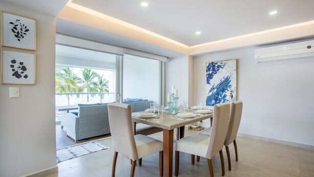 A dining area with a table set for four, a sofa, and a large window with a view of palm trees. - Luxury Beachfront | Corner Condo | Amazing Pools (Puerto Vallarta)