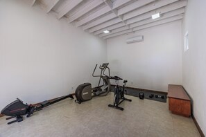 Ein Zimmer mit Deckenventilator, Laufband, Ergometer, einer Holzbank und einem kleinen Schreibtisch.