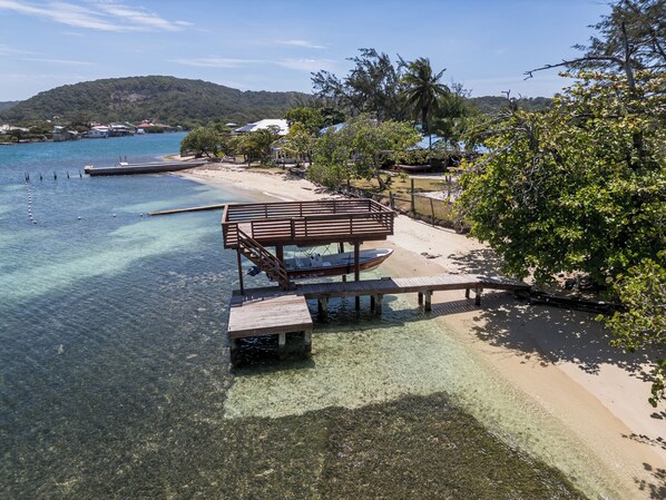 A wooden dock extending into clear blue water, a sandy beach, and a backdrop of hills.