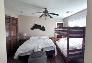 Une chambre avec un lit superposé, un lit simple, un ventilateur de plafond, une lampe fixée au mur et une fenêtre avec des stores.