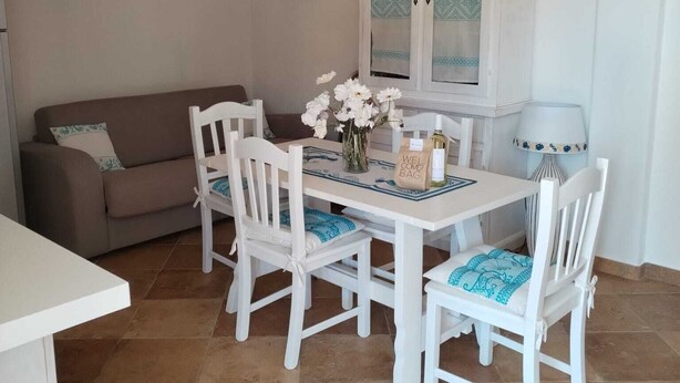 A dining area with a white table, four white chairs, a vase with flowers, a cabinet with glass doors, and a sofa in the background. - "Cozy, newly built villa just 5 minutes from the sea (Sardegna)