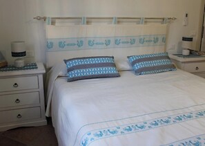 A bed with a white and blue patterned bedspread, two pillows with a blue and white geometric pattern, and a white nightstand with a lamp and a small decorative item. - "Cozy, newly built villa just 5 minutes from the sea (Sardegna)