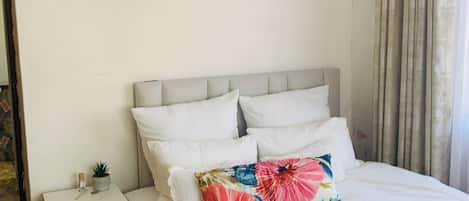 Un dormitorio con cama, mesita de noche, cajón, planta en maceta y ventana con cortinas.