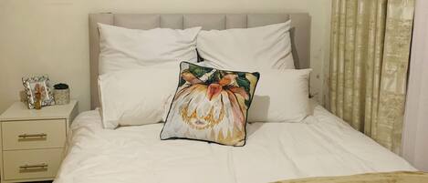 Een net opgemaakt bed met witte lakens en een decoratief kussen, een nachtkastje met een lamp, en een gordijn voor het raam.