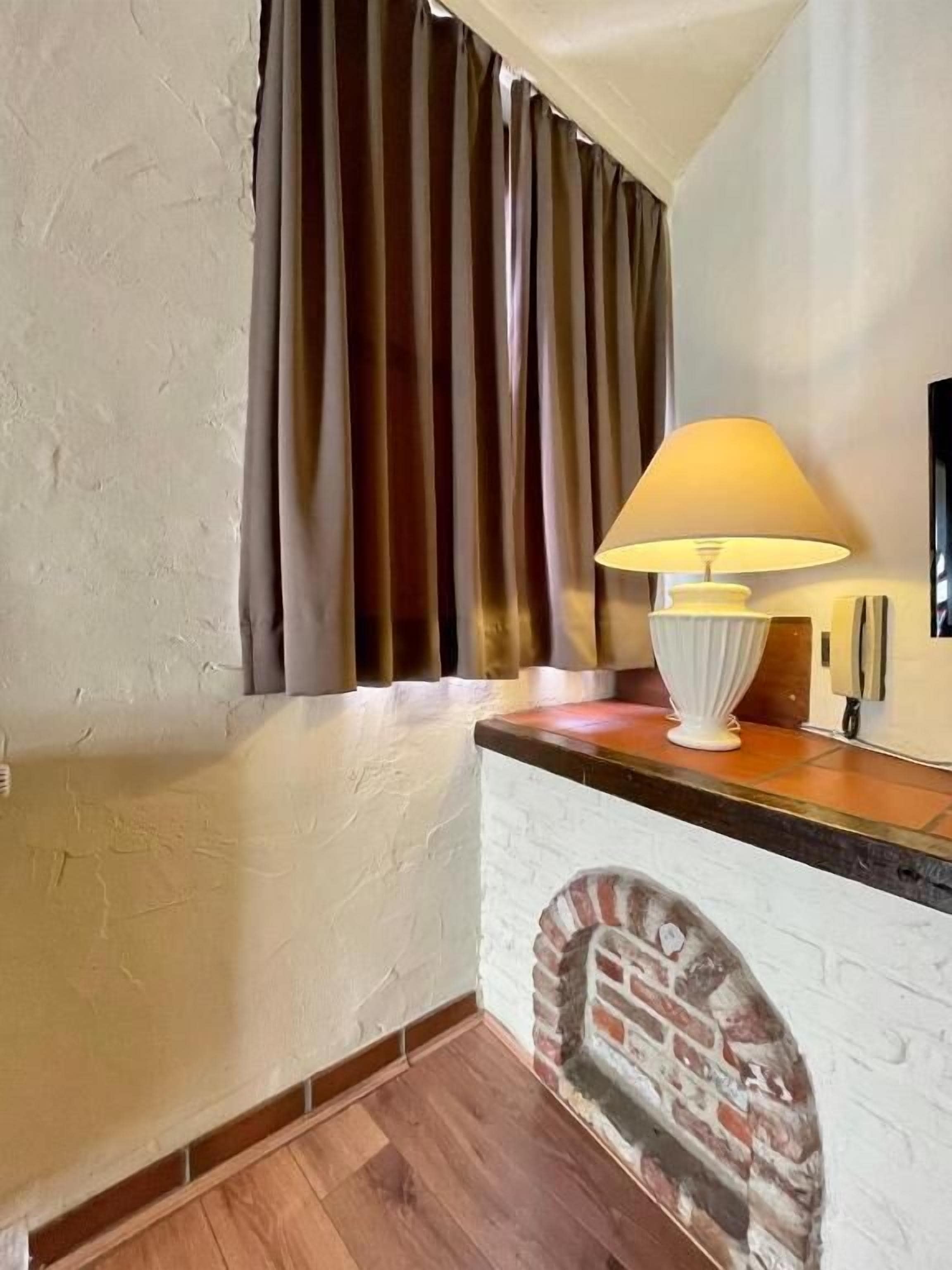 Een kamer met een schouw van bakstenen, een lamp op een houten toonbank en een groot raam met gordijnen.