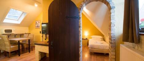 A room with a stone archway, a wooden door, a bed, a desk with a chair, and a skylight.