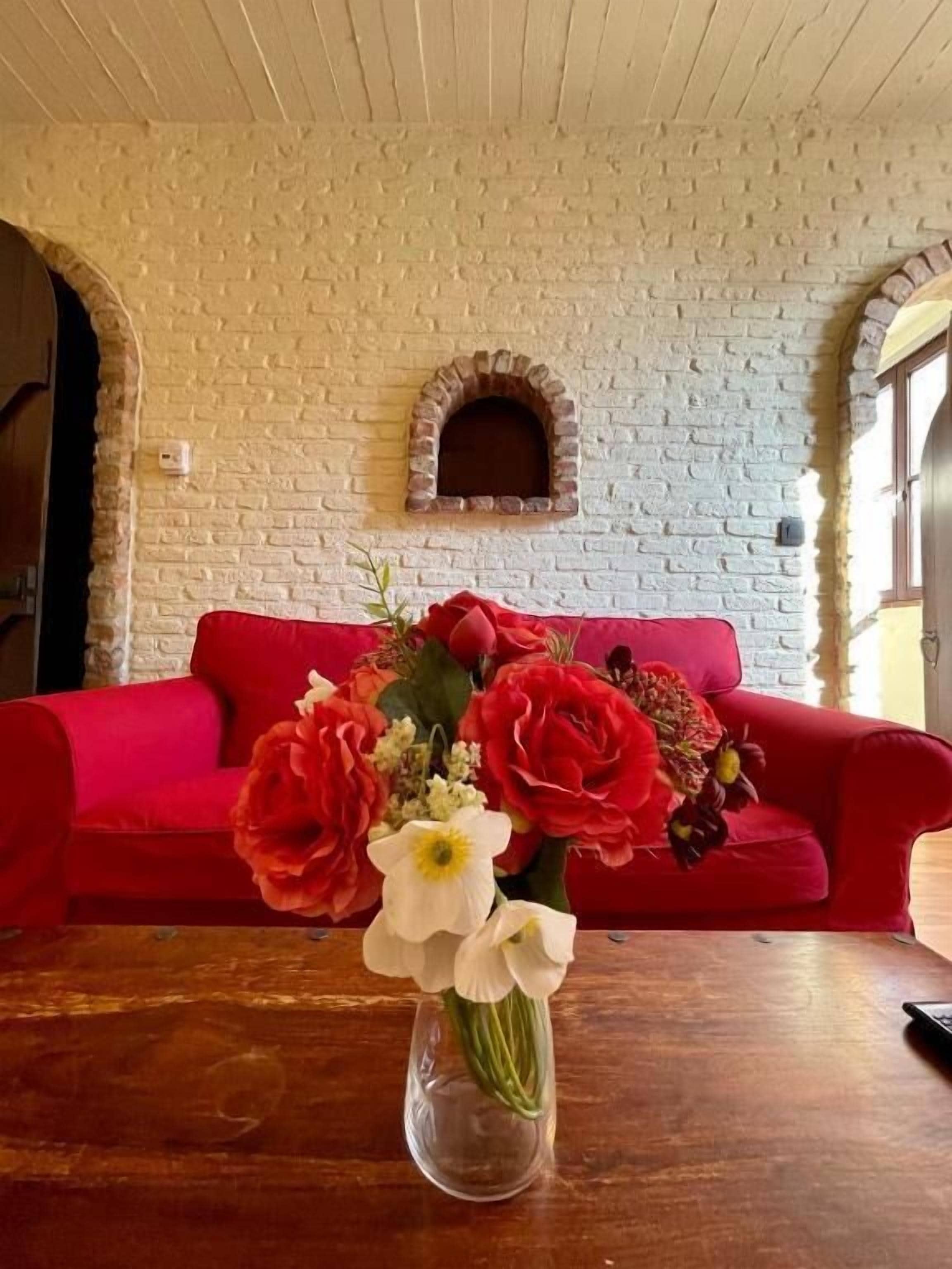 A room with a brick wall, a red sofa, and a vase with flowers on a wooden table.