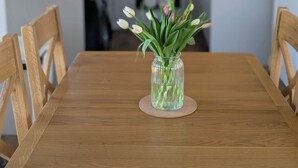 Een eettafel met stoelen, een vaas met bloemen en een keuken op de achtergrond.