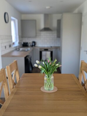 A dining table with chairs, a vase with flowers, and a kitchen in the background. - Culmill Lodges - Birch | Hot Tub Retreat near Loch Ness (Kiltarlity)