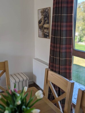 A room with a dining table, chairs, a radiator, a window with plaid curtains, and a painting on the wall. - Culmill Lodges - Birch | Hot Tub Retreat near Loch Ness (Kiltarlity)