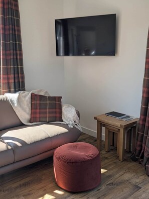 A living room with a sofa, ottoman, and a small wooden table with a tablet. - Culmill Lodges - Birch | Hot Tub Retreat near Loch Ness (Kiltarlity)