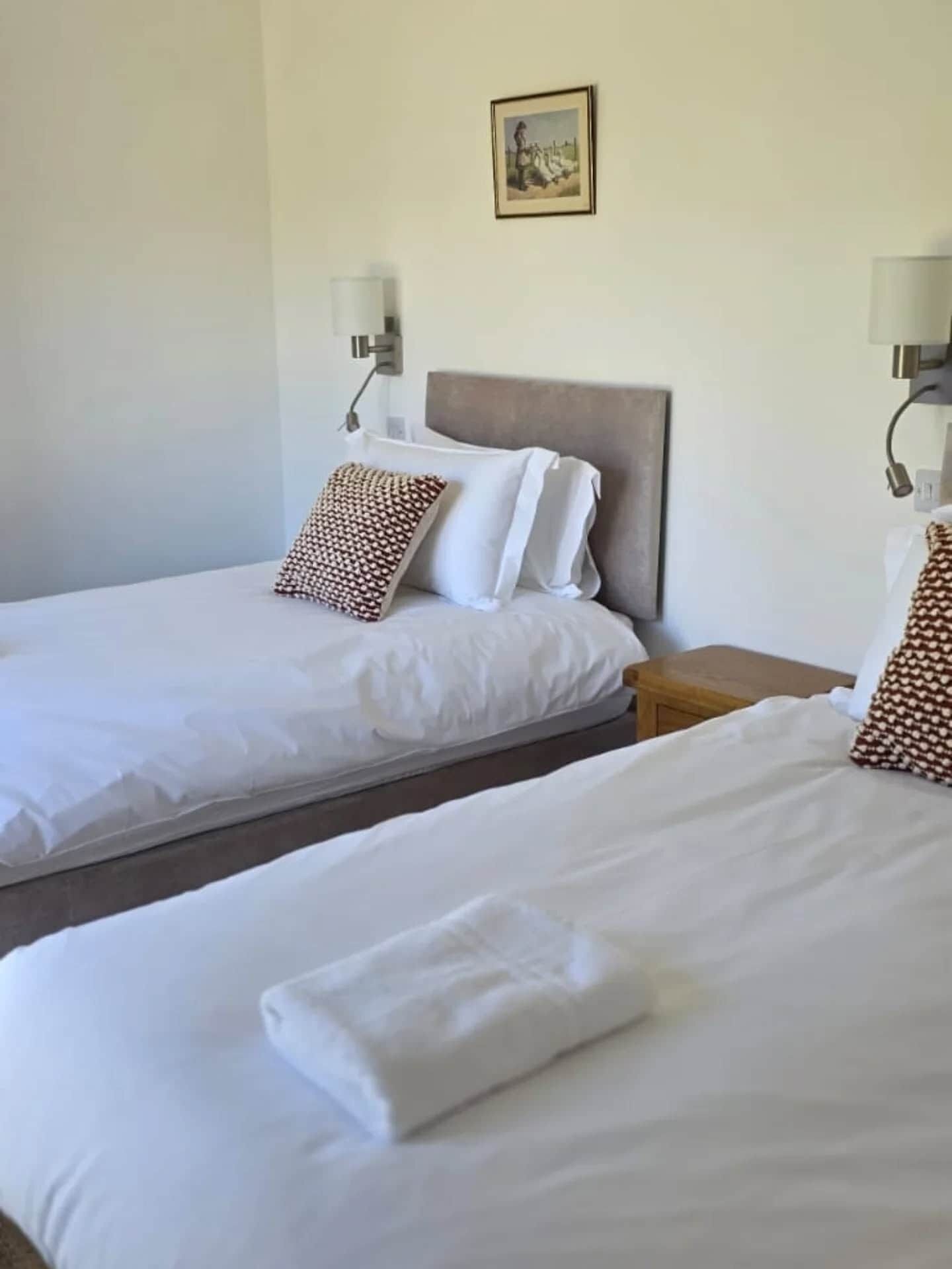 Hotellihuone, jossa on kaksi sänkyä. Jokaisessa sängyssä on valkoiset lakanat ja kuviolliset tyynyt, puinen pääty ja seinällä oleva kehystetty kuva.