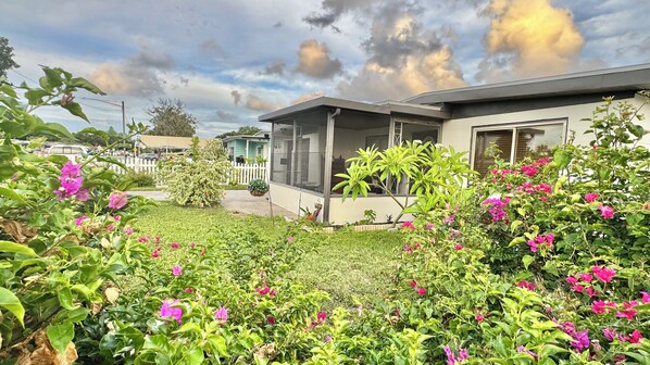 Uma casa com varanda coberta, quintal cercado e diversas plantas floridas.
