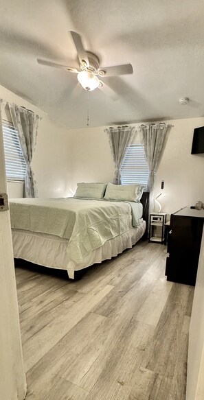 Quarto com cama, ventoinha no teto, mesa de cabeceira, escrivaninha e TV fixada na parede.