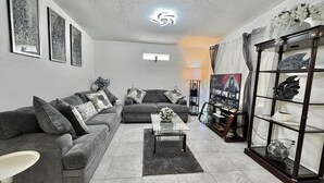 A living room with a grey sofa, a coffee table, a TV on a stand, and a decorative shelf. - Beautifully Renovated Home with Fenced Yard and Gazebo in a Prime Location (Pinellas Park)