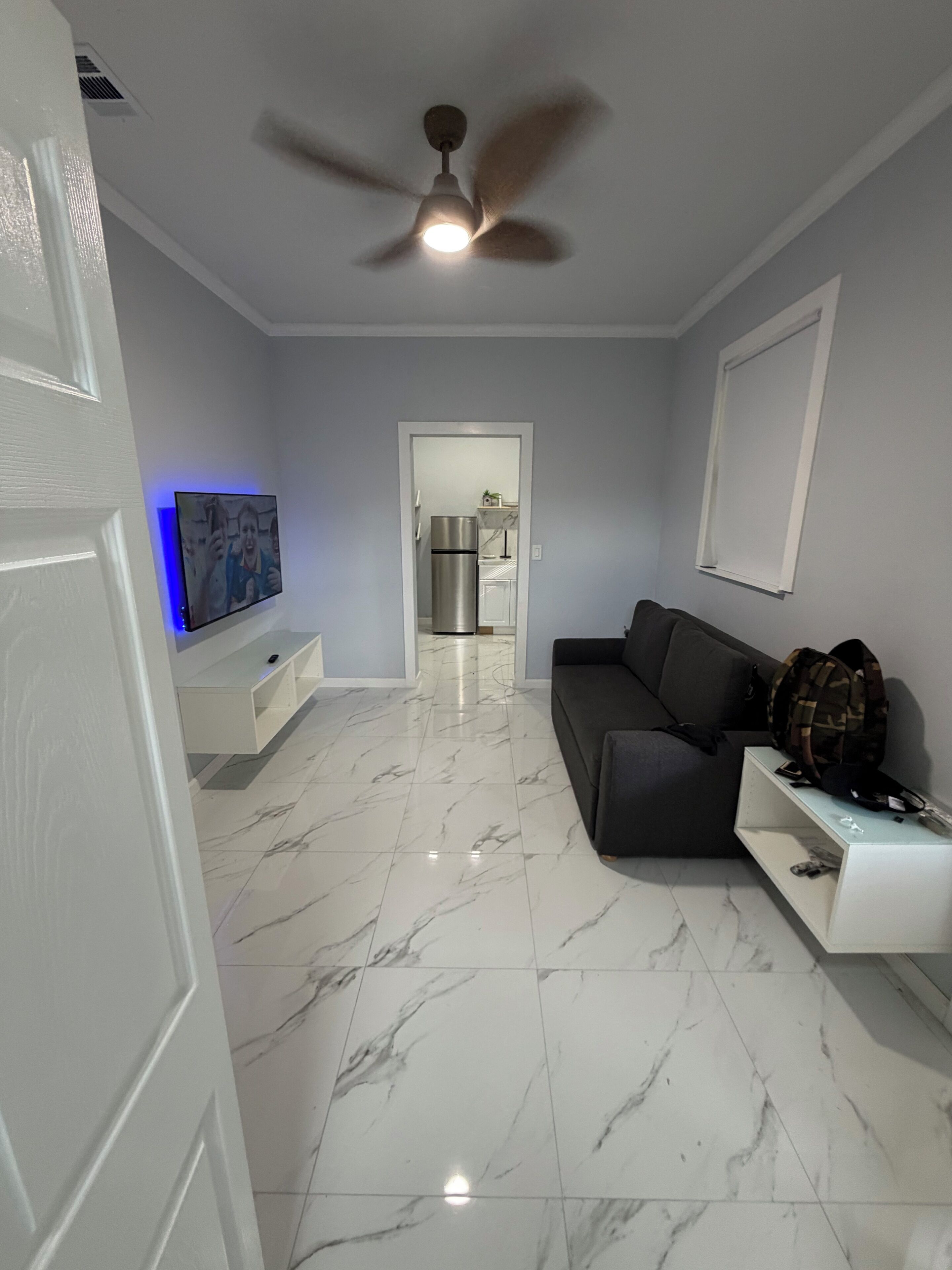 Een moderne woonkamer met een marmeren vloer, een plafondventilator, een flatscreen televisie, een bank en een keuken op de achtergrond.