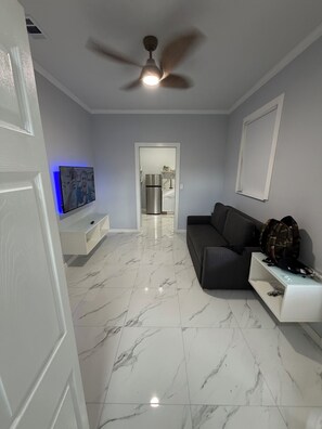 Un salon moderne avec un sol en marbre, un ventilateur de plafond, un téléviseur à écran plat, un canapé et une cuisine en arrière-plan.
