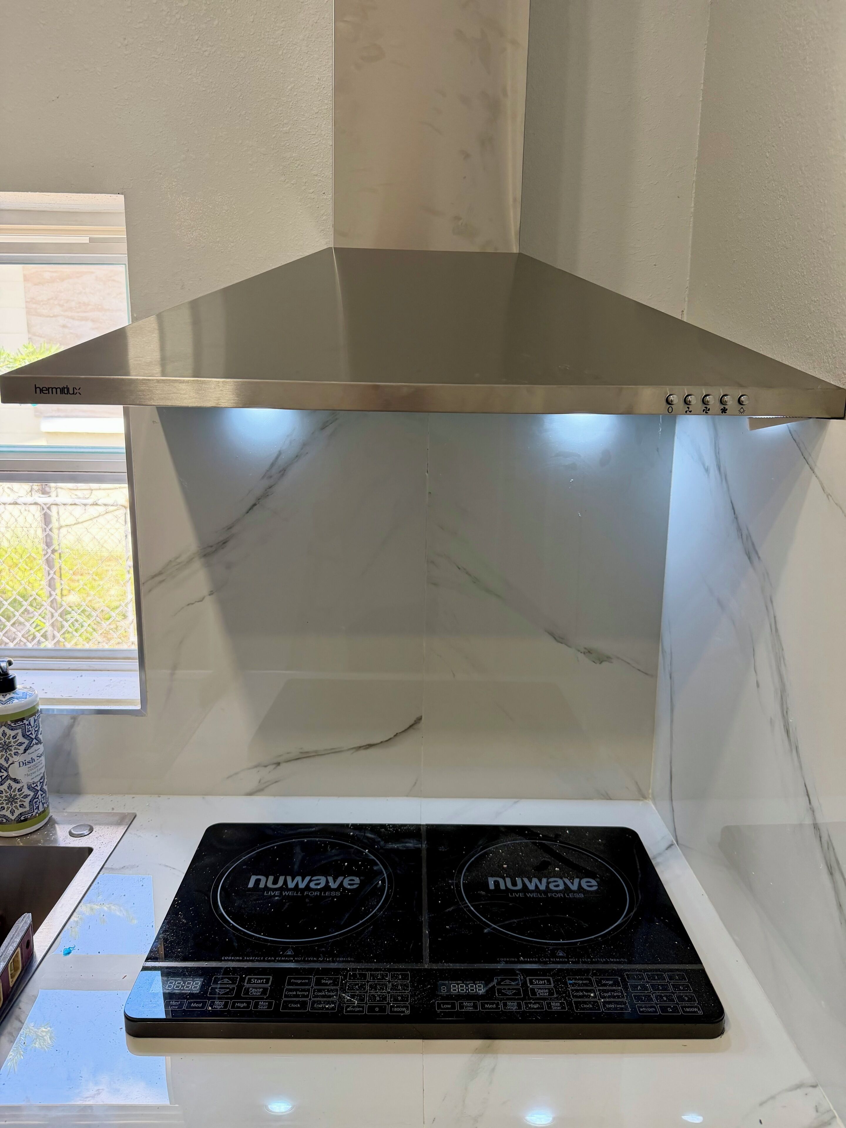 Una cocina con campana extractora de acero inoxidable, dos placas de inducción y un ventanal con vistas a zonas verdes.
