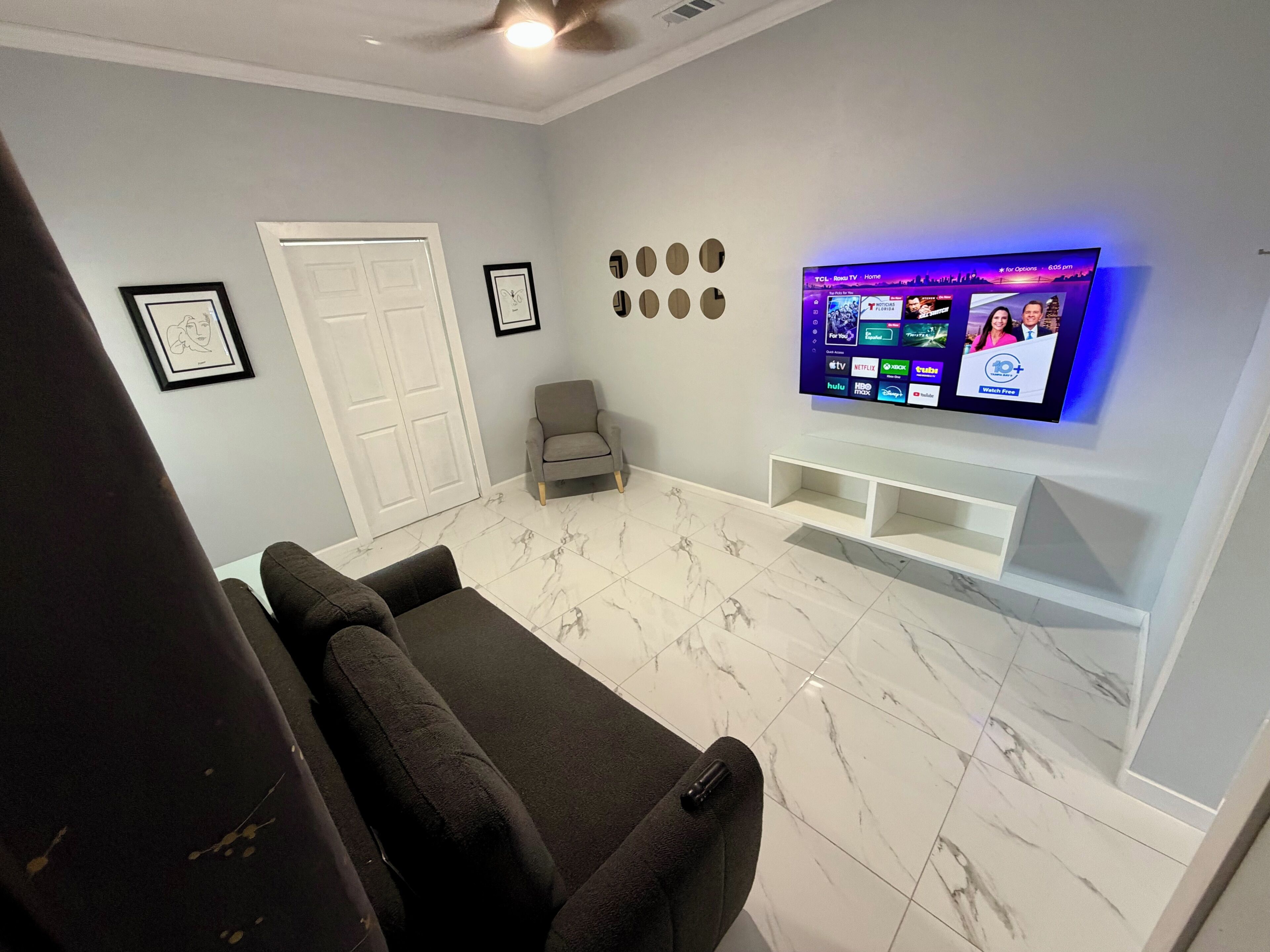 Un salón con una televisión de pantalla plana montada en la pared, un sillón gris y una puerta blanca.