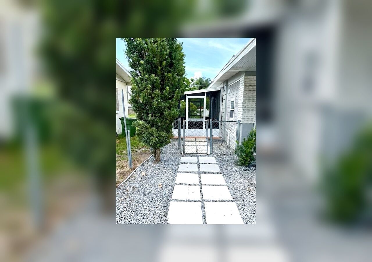 A pathway leading to a building with a covered entrance, surrounded by greenery and a gravel surface.