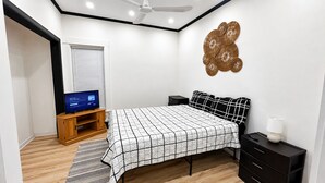 Un dormitorio con cama, televisor sobre un soporte de madera, un adorno en la pared y una mesita de noche con lámpara.