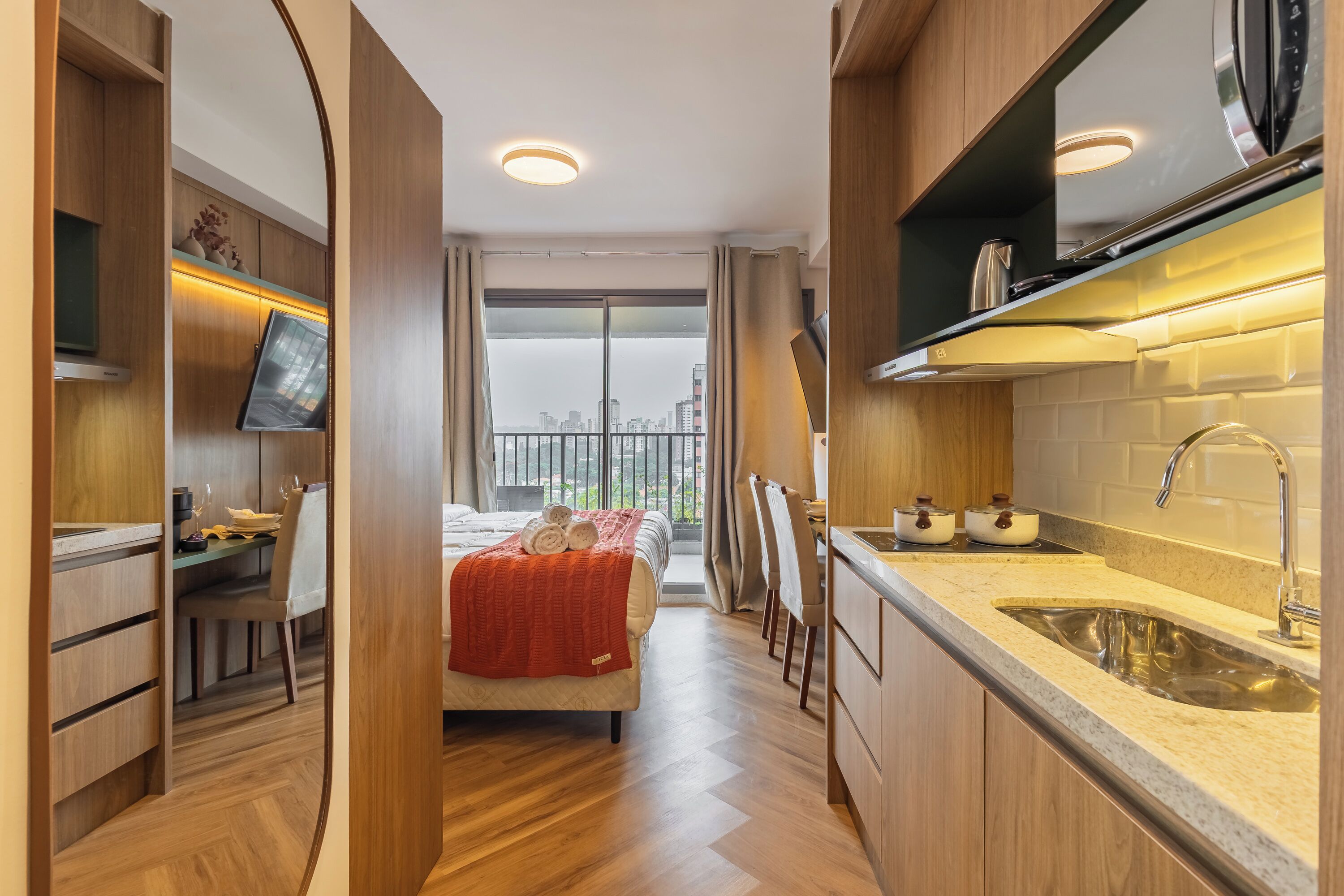 A compact hotel room with a bed, kitchenette, and a balcony with a city view.