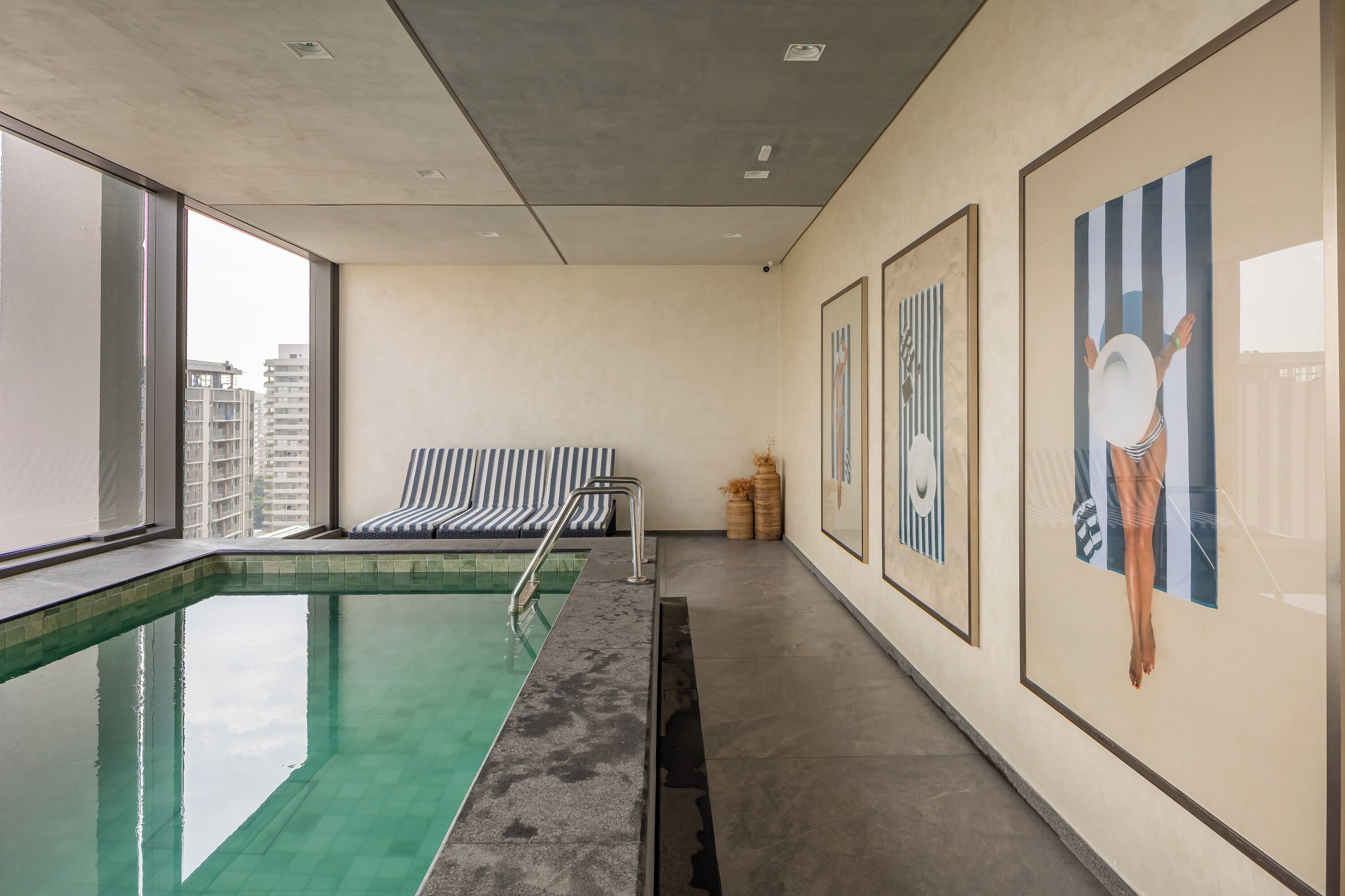 Une piscine avec vue sur les bâtiments, un espace détente avec des chaises à rayures et des œuvres d’art encadrées accrochées aux murs.