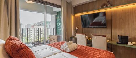 Hotelkamer met een groot bed, een aan de muur bevestigde televisie, een balkon met uitzicht op gebouwen en een koffieapparaat op een bijzettafeltje.