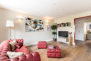 A modern living room with a red sofa, a red ottoman, a flat-screen TV, and a large abstract painting on the wall. - Stylish Bayswater Apartment near Hyde Park (London)