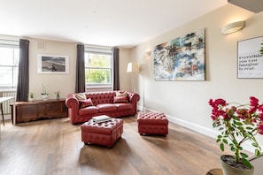 A living room with a red leather sofa, matching ottoman, and a wooden cabinet. There is a painting on the wall and a potted plant on the floor. - Stylish Bayswater Apartment near Hyde Park (London)