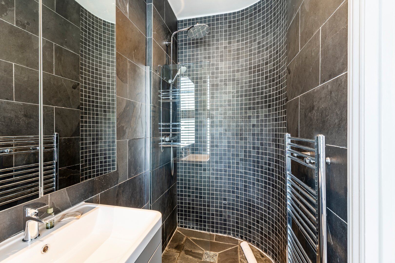 Een moderne badkamer met een gebogen douche, donkere betegelde wanden, een witte badkuip en chroombeslag.