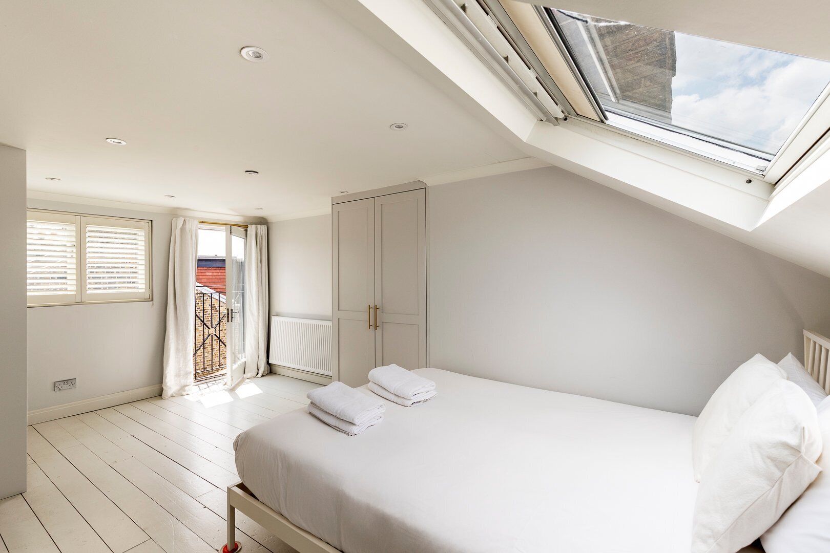A bedroom with a skylight, a bed with white linens, a radiator, and a window with blinds.