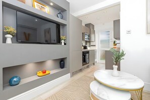 A modern kitchen with built-in appliances, a TV mounted on the wall, and a dining area with a white table and gold legs. - Modern London Apartment for Business Stays (Greater London)