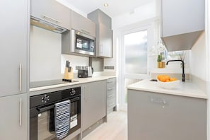 A modern kitchen with grey cabinets, a microwave, a kettle, and a sink with a bowl of oranges. - Modern London Apartment for Business Stays (Greater London)