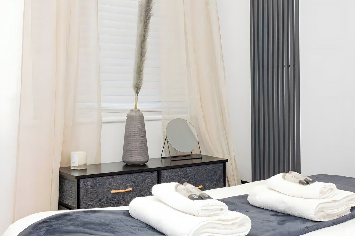 Een slaapkamer met een bed, een dressoir, een spiegel, een vaas met een veer en een raam met gordijnen.