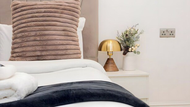 Ein Bett mit beige-farbenem Kopfteil, einer Nachttischlampe, einem weißen Nachttisch und einer Steckdose.