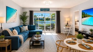 Una sala de estar moderna con un sofá azul, una mesa de centro y un comedor con una mesa redonda y sillas. Se ve una ventana grande con vista a una piscina y palmeras.