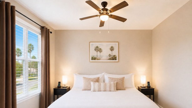 Habitación de hotel con cama, dos mesitas de noche, ventilador de techo, ventana con cortinas e imagen enmarcada en la pared.