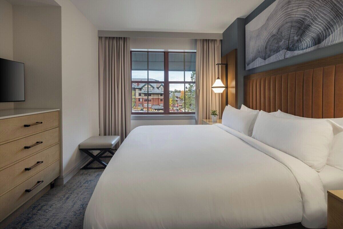 Ein Hotelzimmer mit einem großen Bett, einem Fernseher, einem Schreibtisch mit Schubladen, einem Fenster mit Vorhängen und einem an der Wand befestigten Kunstwerk.