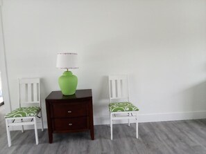 Ein Zimmer mit weißer Wand, einer grünen Vase auf einem Holznachttisch, zwei weißen Stühlen mit grün gemusterten Kissen und einem grauen Teppich.