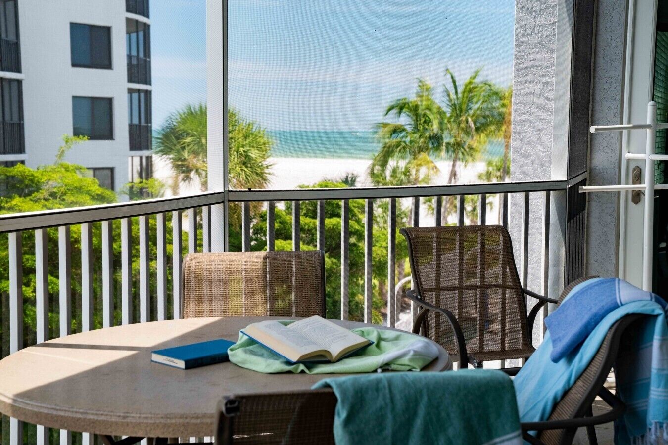A balcony with a table, two chairs, a book, and a view of the beach and palm trees.